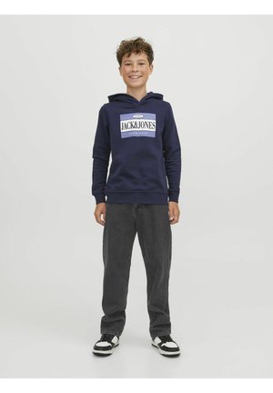 Sweat-shirt à capuche bleu marine avec logo sur le devant, associé à un jean noir et des baskets noir et blanc, se tenant devant un fond uni.