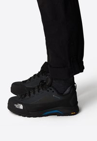 Svarta vandringsskor med en texturerad ovandel, snörningssystem och en sula i blått och gult som har Vibram-teknologi.