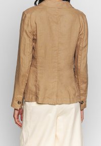 Giacca di lino beige con un design aderente, colletto a revers, maniche lunghe e polsini con bottoni. Cuciture semplici e texture liscia visibili.