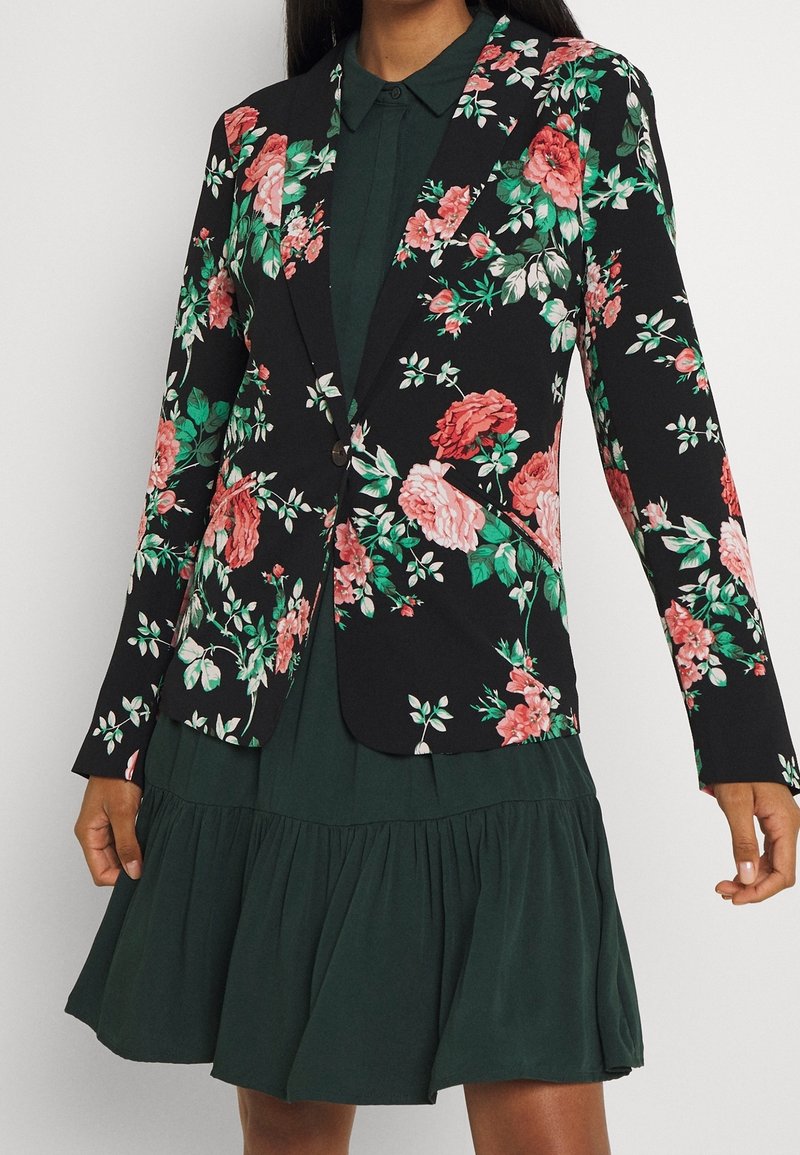 Mujer vistiendo un blazer negro con estampado floral en tonos rosa y verde sobre un vestido verde oscuro con cuello y falda de capas.