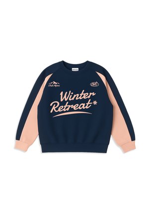 Young man with curly blond hair wears navy and light pink sweatshirt with text "Winter Retreat" and white pants, hands in pockets.