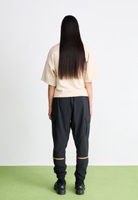 Person with long straight dark hair wearing beige short-sleeve shirt, black cargo pants with tan stripes, and black sneakers, standing on green floor.