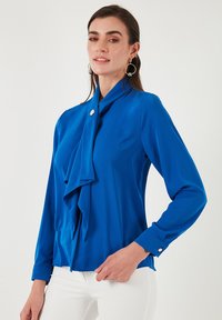 Bluză albastră cu mânecă lungă, realizată dintr-un material fin, având un design cu un front drapat și o guler cu detalii decorative sub formă de nasturi.