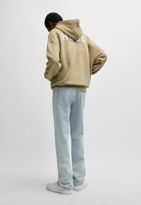 Khaki hoodie met witte logo en tekst op de achterkant, gecombineerd met lichtblauwe geruite broek en witte sneakers. Casual outfit, relaxte pasvorm.