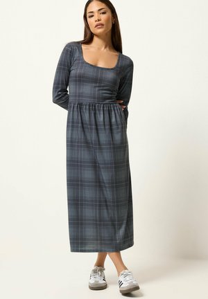 Kobieta stojąca ubrana w długą szarą kraciastą sukienkę z długimi rękawami i białe trampki na jednolitym jasnym tle.