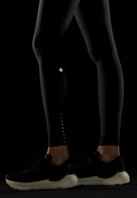Leggings negros con material texturizado y detalles reflectantes en el tobillo, combinados con zapatillas deportivas negras que tienen una suela blanca.