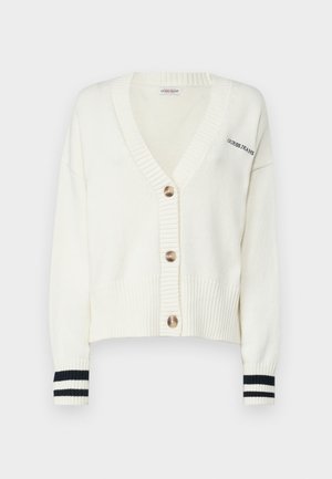 Valkoinen neulecardigan V-pääntiellä, jossa on neljä suurta nappia ja musta-valkoiset raidat hihoissa. Tekstissä lukee "GUESS JEANS."