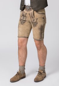 Beige Lederhosen mit aufwendiger dunkler Stickerei, Seitentaschen und Knopfdetails. Kombiniert mit braunen Schuhen und grauen gestrickten Socken.