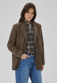 Sacou maro din suede cu un singur nasture, revere croite, și două buzunare frontale, asortat cu o cămașă cu carouri și blugi albaștri.