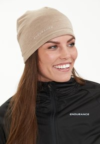 Beige texturerad mössa med en åtsittande design, prydd med "ENDURANCE"-logotyp i vitt. Matchad med en svart zip-up jacka.