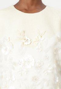 White textured garment featuring 3D floral appliqués and beaded accents, with a rounded neckline and intricate detailing throughout.