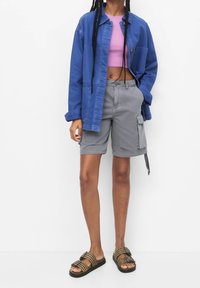 Niebieska kurtka dżinsowa, fioletowy krótki top, szare szorty cargo z kieszeniami oraz czarne sandały z brązowymi paskami. Prosty, codzienny strój.