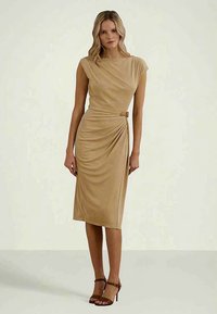 Mujer de pie con un vestido sin mangas, de longitud a la rodilla y color beige, con pliegues laterales y detalle de hebilla, que lleva sandalias de tacón alto marrones.