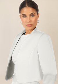 Biała, dopasowana marynarka z wysokim kołnierzem, strukturalnymi ramionami i gładką fakturą. W zestawieniu z białą bluzką wnętrzową i minimalnym dodatkiem.