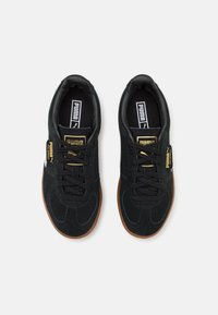 Tenisi negri din suede, cu vârf rotunjit, șireturi negre și talpă din cauciuc colorat. Prezintă un etichetă cu logo aurie și elemente de design texturate.