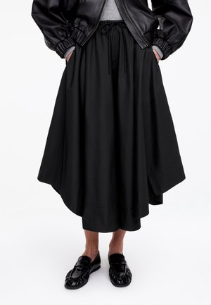 Personne portant une veste en cuir noire, un haut gris, une jupe midi asymétrique noire avec taille à cordon, et des mocassins noirs, debout les mains dans les poches.