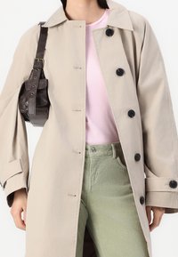 Personne portant un trench-coat beige à boutons sur une chemise rose et un pantalon vert, portant un sac à bandoulière en cuir marron foncé.