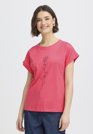 Femme souriante aux cheveux bruns courts portant un T-shirt rose aux manches retroussées avec un dessin au trait noir d'une rose et un jean bleu.