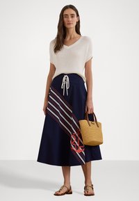 Σκούρα μπλε φούστα με διαγώνιες λευκές και κόκκινες ρίγες, με σχέδιο άγκυρας; συνδυασμένη με ένα ελαφρύ, κρεμ τοπ και μια υφασμένη τσάντα από ψάθα.
