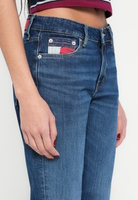 Traperice od trapera u srednjoj plavoj boji s pohabanom teksturom, s printanim dizajnom džepa u crvenim, bijelim i plavim naglascima, u standardnom kroju.