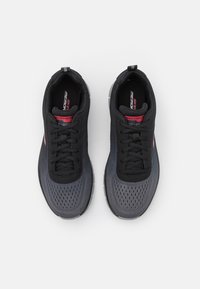 Black and grey athletic shoes with a mesh upper, round toe, laced design, and contrasting red accents on the tongue and side logo.