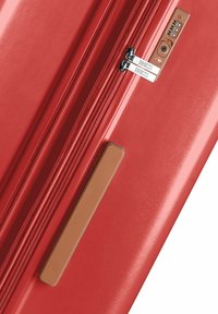 Maleta rígida roja con una superficie lisa, dobles cremalleras, cerradura de combinación y un mango de agarre texturizado marrón.