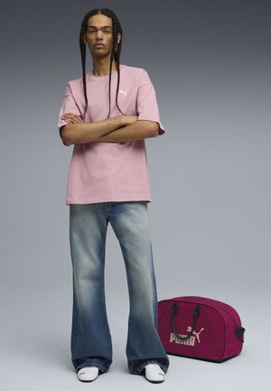 Lys pink bomulds t-shirt, løse slidte blå jeans og hvide sneakers. Mørkerød duffel taske med sorte detaljer og "PUMA" branding på siden.