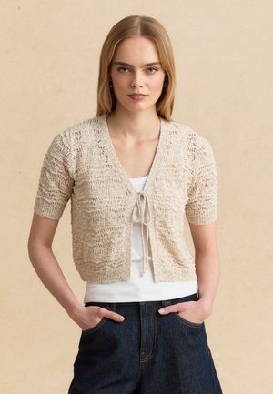 Femme aux cheveux blonds raides portant un cardigan beige en maille à manches courtes par-dessus un top blanc et un jean en denim foncé, les mains dans les poches.