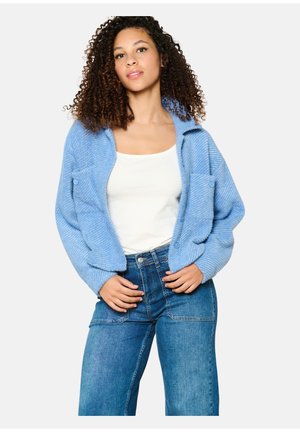 Modrý pletený cardigan s texturovaným vzorem, oversize střih a dvě přední kapsy. Nesený přes bílý top a spárovaný s vysokými džínami.