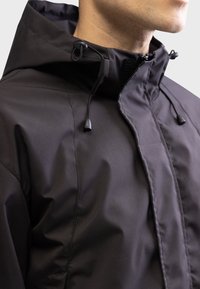 Zwarte waterproof jack met hoge kraag, verstelbare capuchon met trekkoorden en een gladde matte textuur. Rits aan de voorkant.