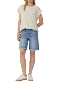 Lichtbeige, kort mouwtop met een stergrafiek, gecombineerd met opgerolde mouwen, middelblauwe spijkershorts en witte sneakers met beige accenten.