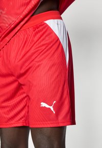 Vörös atlétikai rövidnadrág könnyű anyagból, fehér oldalsó panellel és a szegély közelében található fehér Puma logóval. Texturált felületű dizájn.