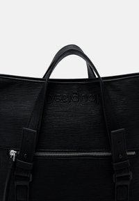 Czarna torebka ze skóry z fakturą, z dwoma uchwytami, przednią kieszenią na zamek i markowym detalem. Posiada zwartą formę i solidne paski.