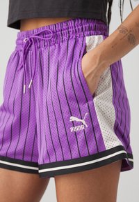 Short en mesh violet avec des rayures verticales noires, des panneaux latéraux blancs, un ourlet noir et blanc, une taille élastique et une main dans la poche droite.