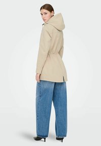 Veste à capuche beige avec taille élastique, associée à un jean denim bleu clair surdimensionné. Tissu lisse avec une coupe décontractée et des fentes sur les côtés.