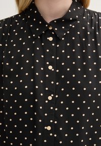 Camisa negra con un frente de botones, que presenta un patrón de lunares en beige claro, cuello clásico y una textura de tela suave y lisa.