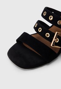 Μαύρο σουέτ σανδάλι με δύο φαρδιές λουρίδες που διαθέτουν χρυσούς κρίκους και τετράγωνη μύτη. Η ρυθμιζόμενη αγκράφα προσθέτει λεπτομέρεια στο σχέδιο.