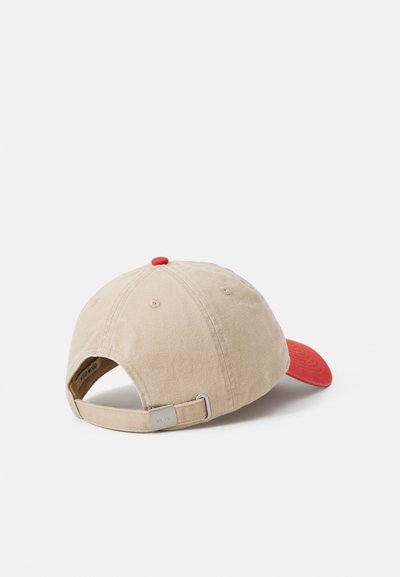 Baseball cap i beige bomull med en rød skjerm, med en stoffreim og metalljusteringsspenne bak. Strukturert stofffinish.