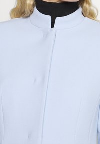 Donna che indossa un cappotto blu chiaro con colletto rialzato sopra un maglione a dolce vita nero, che mostra la parte superiore del petto e del collo.