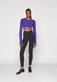 Lilla langermet crop-top med "NIKE PRO" branding, kombinert med høyhalsede svarte leggings og svarte treningssko.