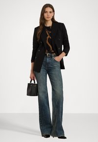 Svart dobbelspent blazer over en mønstret skjorte, kombinert med vide blå jeans og holder en liten svart veske.