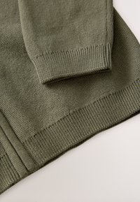 Manche et ourlet d'un pull en maille vert olive avec poignets et bande côtes sur une surface claire.