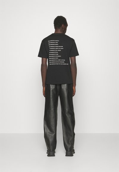 Férfi fekete pólót visel, fehér szöveggel a hátán, és fekete bőr nadrágot, egy sima fehér fal felé állva.