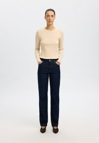 Beige gerippter Langarmoberteil, dunkelblaue gerade Jeans, Hände in den Taschen, braune Schuhe. Einfaches, lässiges Outfit ohne Muster.
