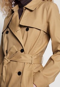 Gros plan d'une personne portant un trench beige avec des boutons noirs, une ceinture nouée et un col roulé marron en dessous.