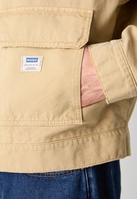 Beżowa kurtka z płótna z kieszenią na piersi i metką. Charakteryzuje się mocnym szwem, prostym krojem i gładką teksturą. Noszona na dżinsy.