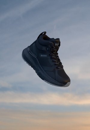 Basket montante noire avec lacets et logo BOSS flottant devant un ciel nuageux au coucher du soleil.