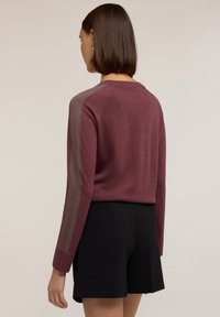 Burgundin villapaita pitkillä hihoilla, joissa on kontrastivärinen tummempi ruskea raita jokaisessa hihassa, yhdistettynä mustiin shortseihin.