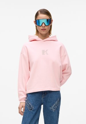 Oseba, ki nosi prevelika modra ogledalna sončna očala, svetlo roza pulover s kapuco z logotipom in modre jeans hlače s srebrnimi vezalkami na stegnih.