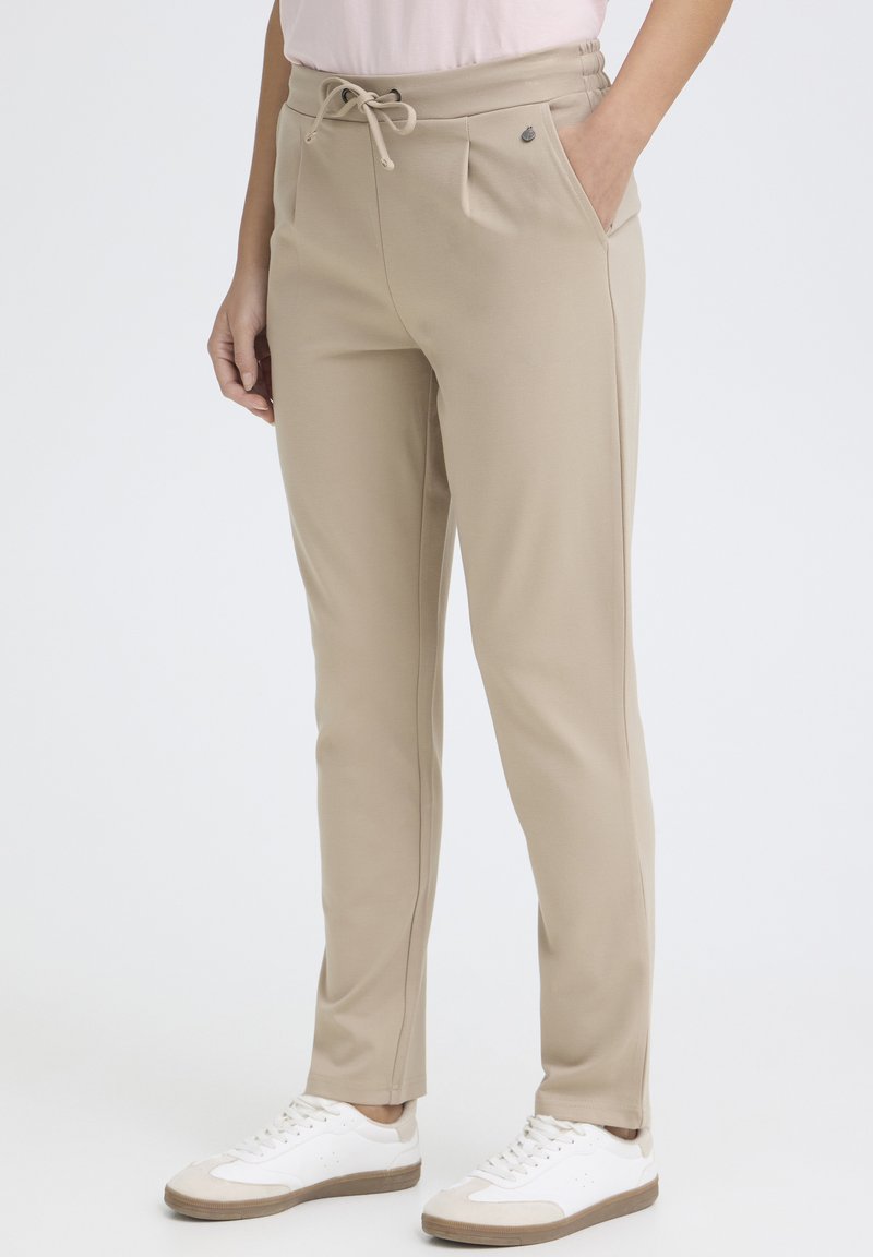 Bézs színű, zsinóros, hétköznapi nadrág fehér tornacipővel, zsebek láthatóak, a modell világos, egyszínű háttér előtt áll.